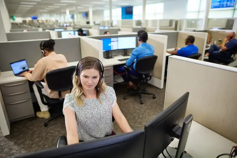 A woman wearing a headset works at a computer in a busy professional call center with other employees in cubicles.
